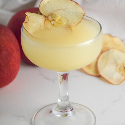 Small cocktail glass with filled with golden cocktail and garnished with dried apple slice on a white background with red apple in background