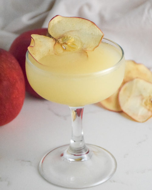 Small cocktail glass with filled with golden cocktail and garnished with dried apple slice on a white background with red apple in background