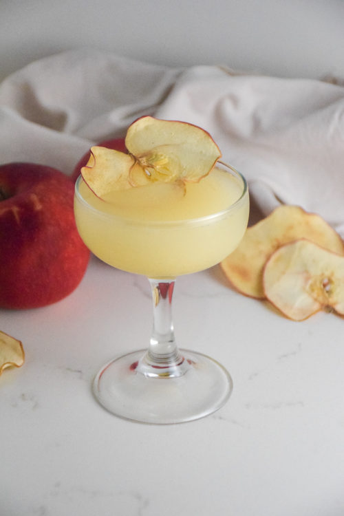 Small cocktail glass with filled with golden cocktail and garnished with dried apple slice on a white background with red apple in background