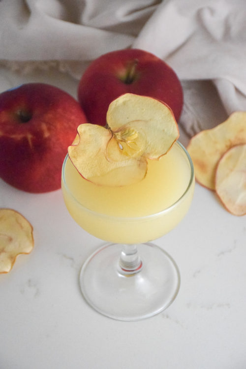 Small cocktail glass with filled with golden cocktail and garnished with dried apple slice on a white background with red apple in background