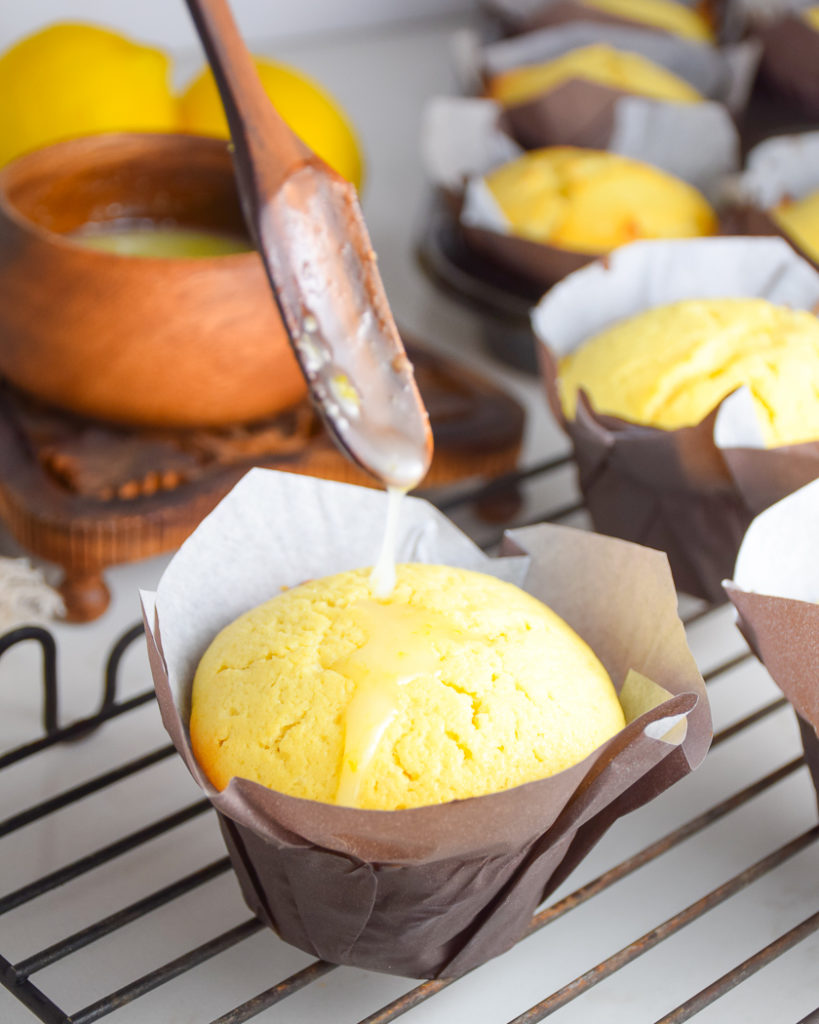 yellow lemon cakes in paper muffin wrappers on metal cooling rack with brown wooden spoon drizzling glaze