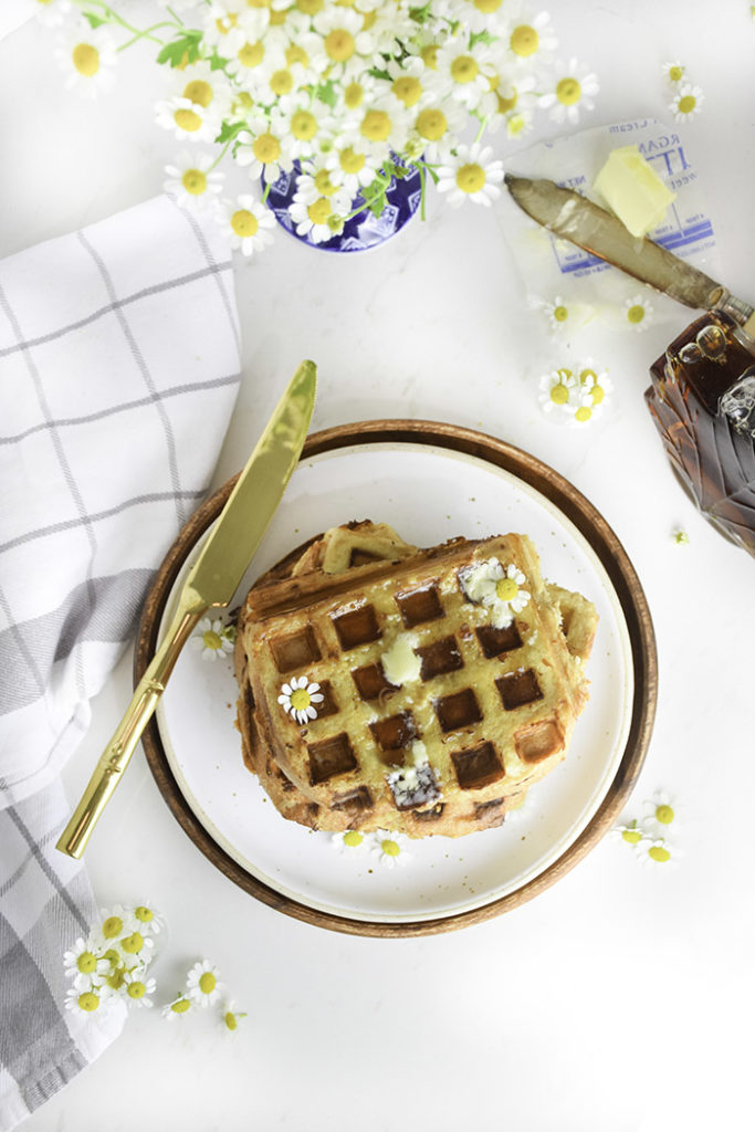 french toast waffles on a white plate with gold knife on edge of plate and folded kitchen towel, chamomile flowers scattered around 
