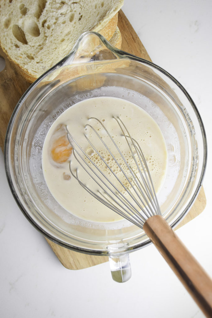 glass mixing bowl with whisk and eggs and milk in bowl on a wooden cutting board