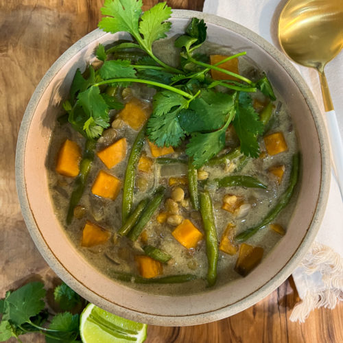 Soup in a pink bowl with orange sweet potato chunks, green beans and bright green cilantro stems on top on a wooden background with napkin and spoon on the right side