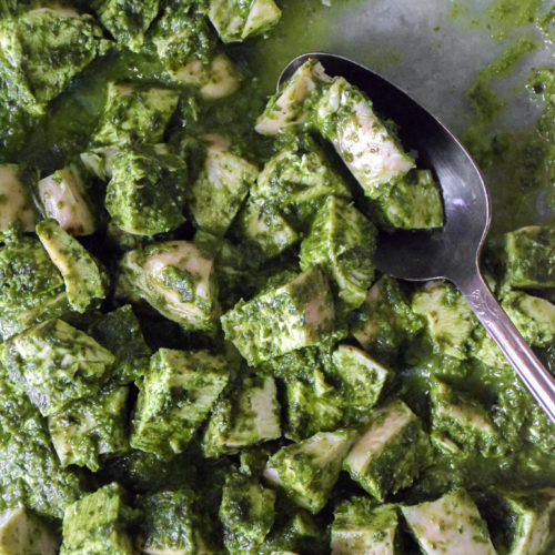 Bite sized cubes of chicken breast covered in a green pesto sauce in a clear baking dish with a spoon