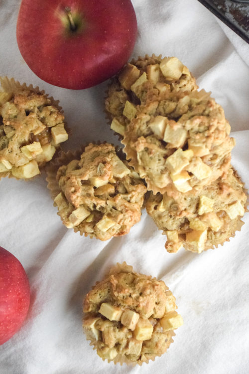 Muffin with chunks of apple on a white background with red apple and muffins in the background