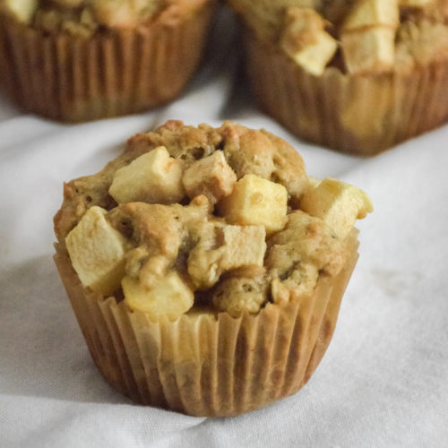 Muffin with chunks of apple on a white background with red apple and muffins in the background