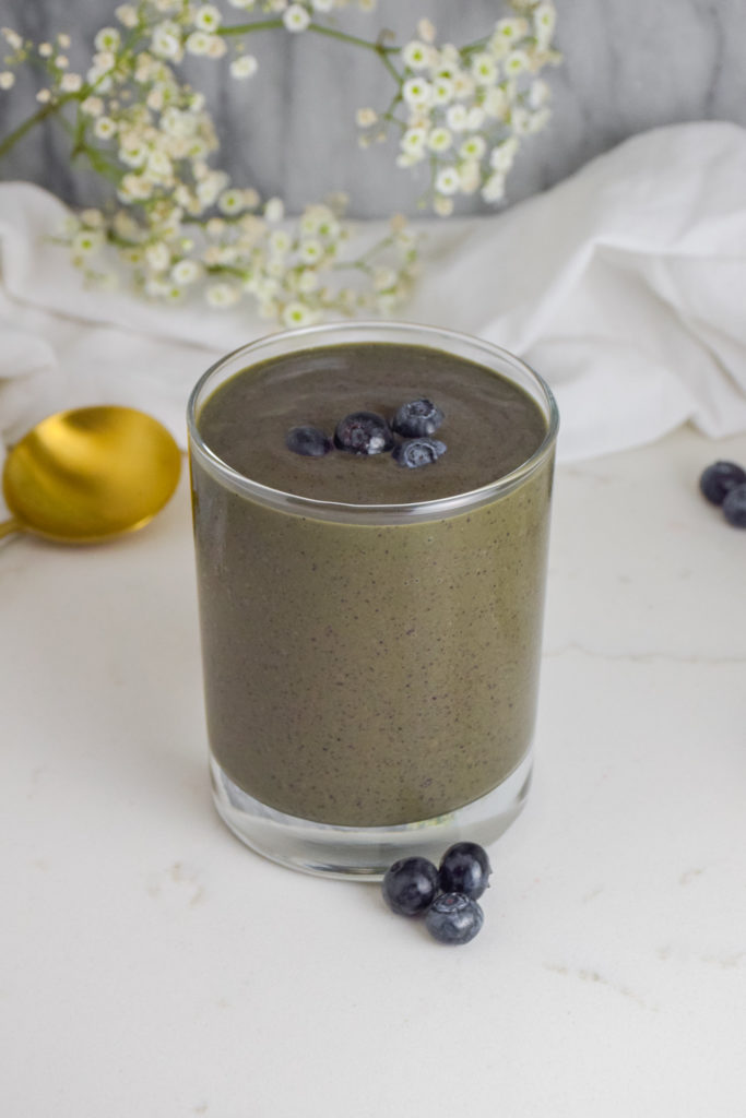Blue smoothie in a clear glass with fresh blueberries on top. Marble background with flowers