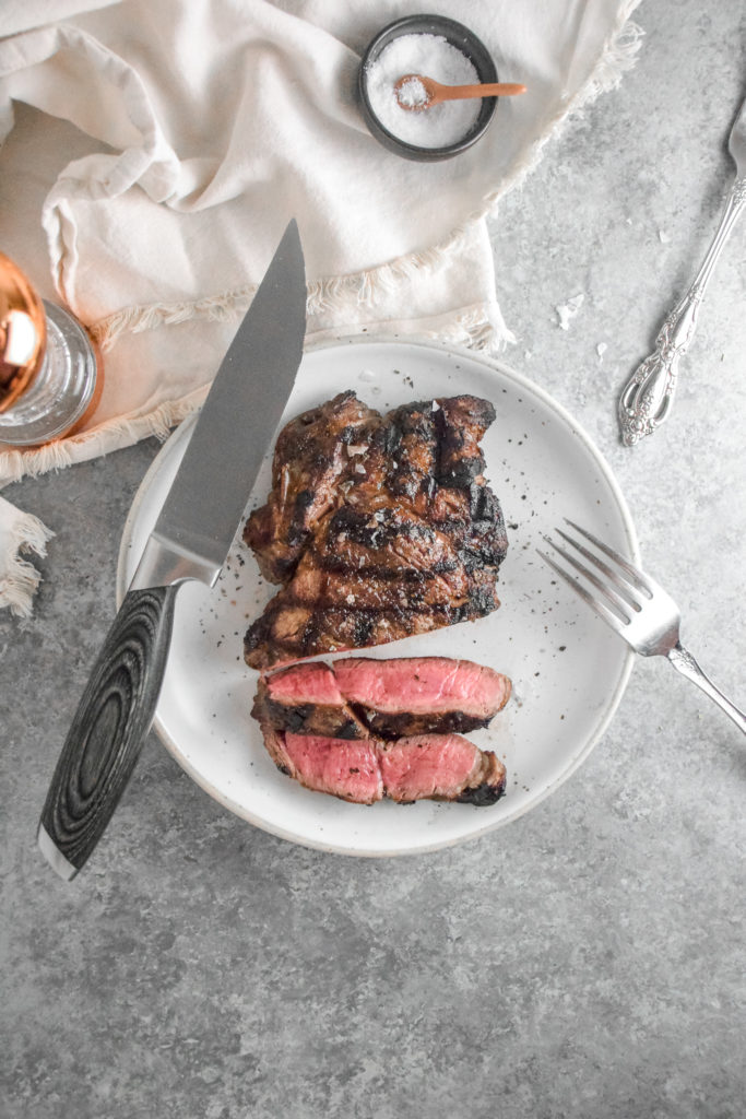 Ribeye Steak with criss cross grill marks cooked to medium rare pink inside on a white plate with grey background and silver fork on the right