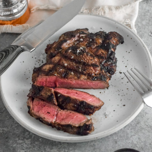 Ribeye Steak with criss cross grill marks cooked to medium rare pink inside on a white plate with grey background and silver fork on the right