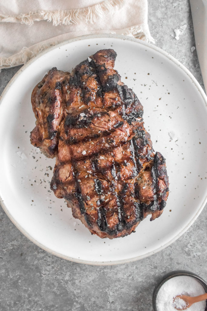 Ribeye Steak with criss cross grill marks cooked to medium rare pink inside on a white plate with grey background and silver fork on the right