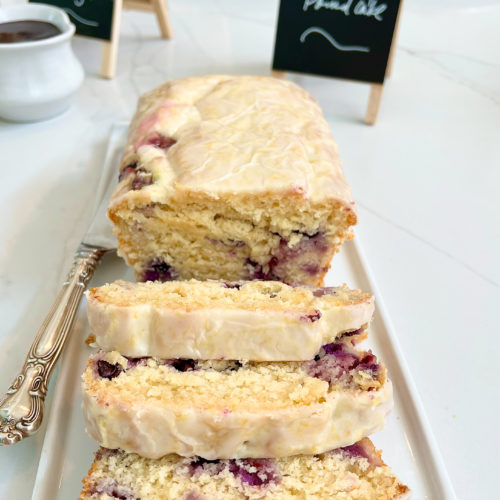 Lemon Blueberry Pound Cake with lemon glaze. Three slices folded over in the front of image and cake in the background on a white plate with black mini chalkboard in the background