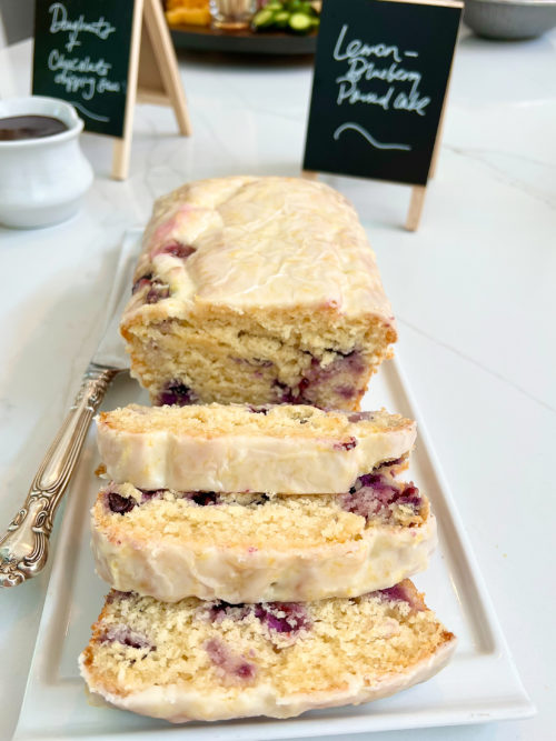 Lemon Blueberry Pound Cake with lemon glaze. Three slices folded over in the front of image and cake in the background on a white plate with black mini chalkboard in the background