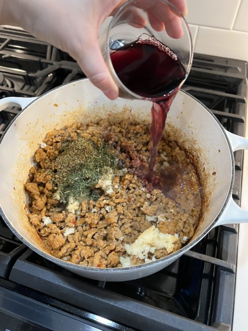 meat sauce in a white pot on stove with dried herbs and red wine being poured in