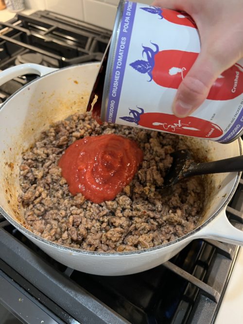 meat sauce in a white pot on stove with can of crushed tomatoes being poured in