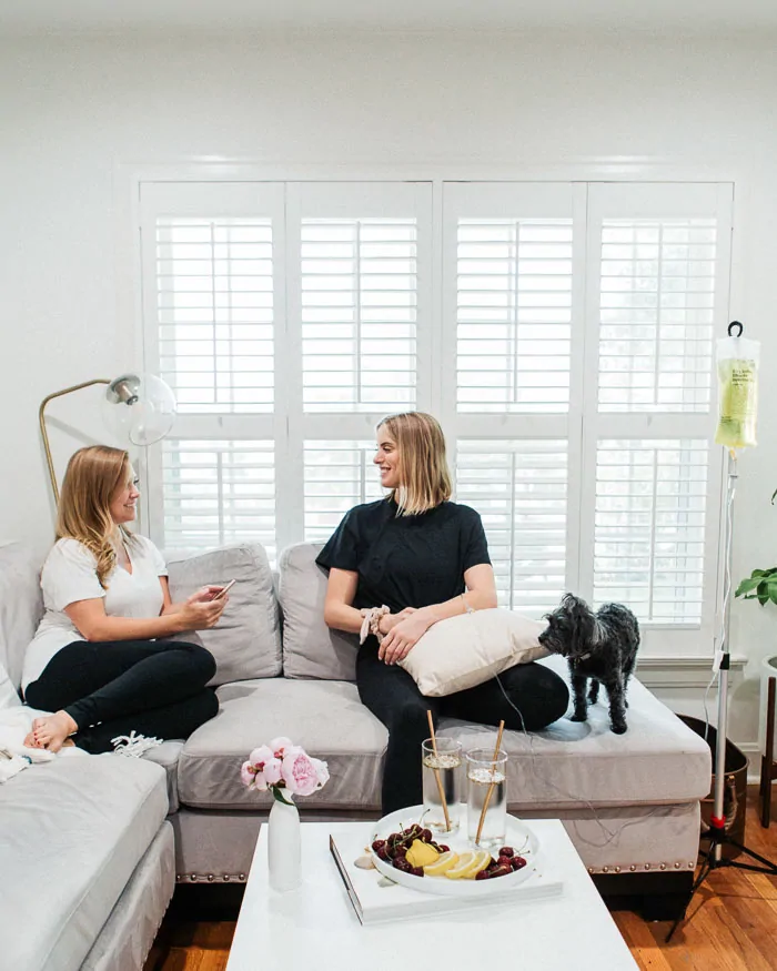 Two women sitting on couch, woman on right is getting an IV drip and there is a black dog on the sofa