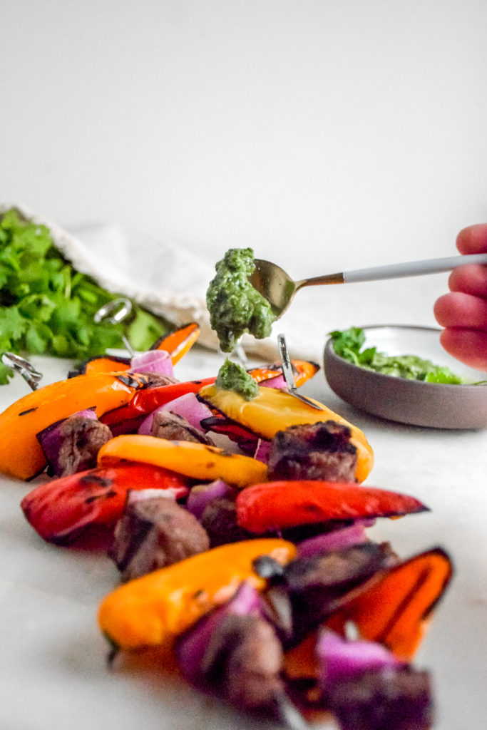 Sirloin Steak with red and orange baby bell peppers and red onion on metal skewers with black grill marks on a white background and green cilantro chimichurri being drizzled on top