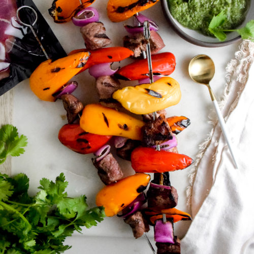 Sirloin Steak with red and orange baby bell peppers and red onion on metal skewers with black grill marks on a white background