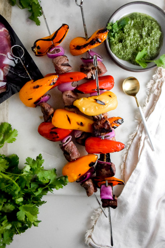 Sirloin Steak with red and orange baby bell peppers and red onion on metal skewers with black grill marks on a white background