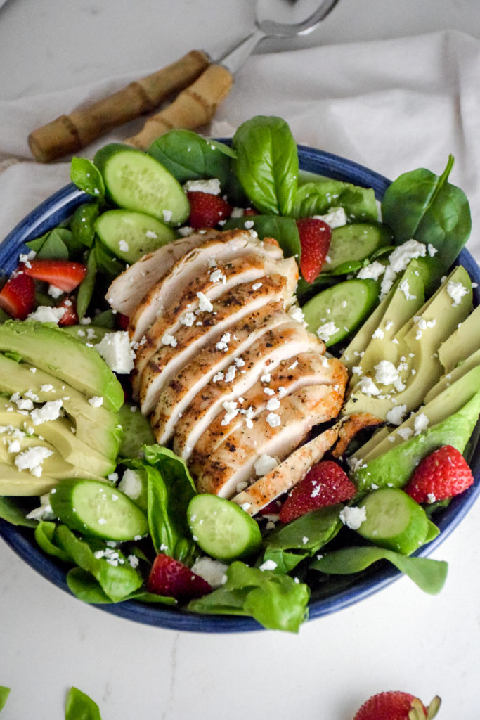 Salad with sliced grilled chicken breast in the middle and avocado and strawberries and feta cheese on top all on a white background