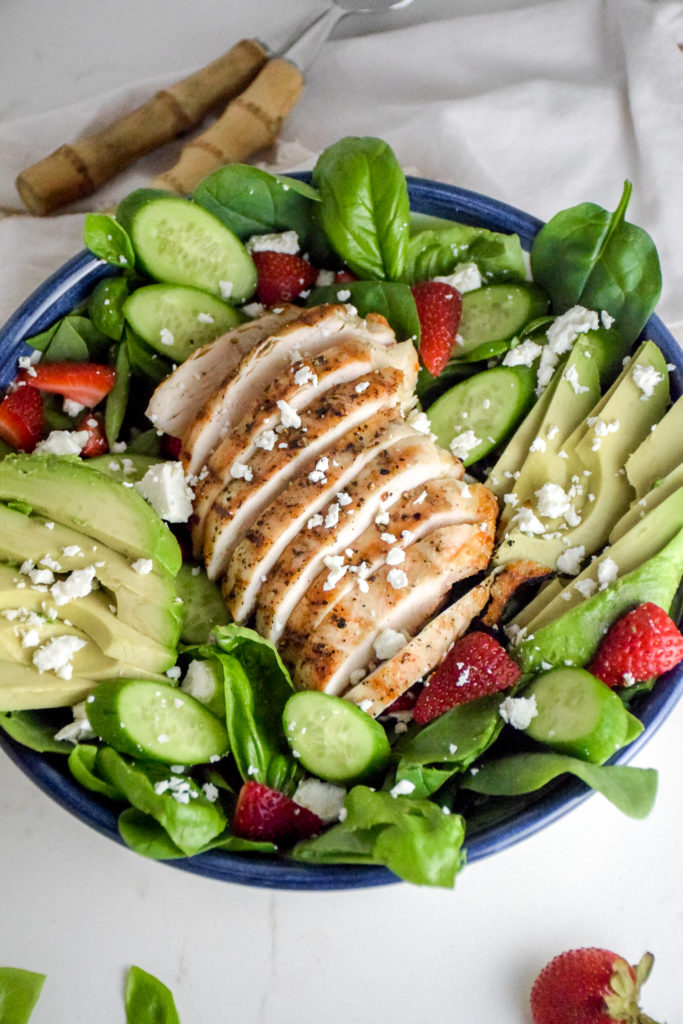 Salad with sliced grilled chicken breast in the middle and avocado and strawberries and feta cheese on top all on a white background