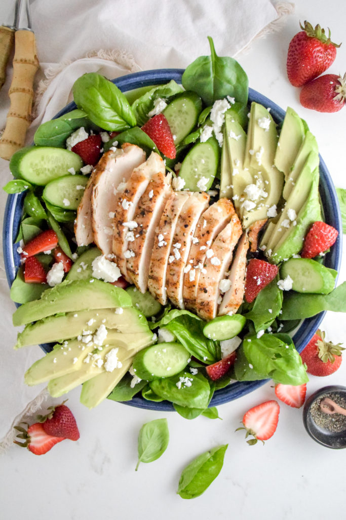 Salad with sliced grilled chicken breast in the middle and avocado and strawberries and feta cheese on top all on a white background