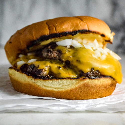 Burger with two thin patties on a bun with cheese and onions on a crumbled white paper on the grey marble background