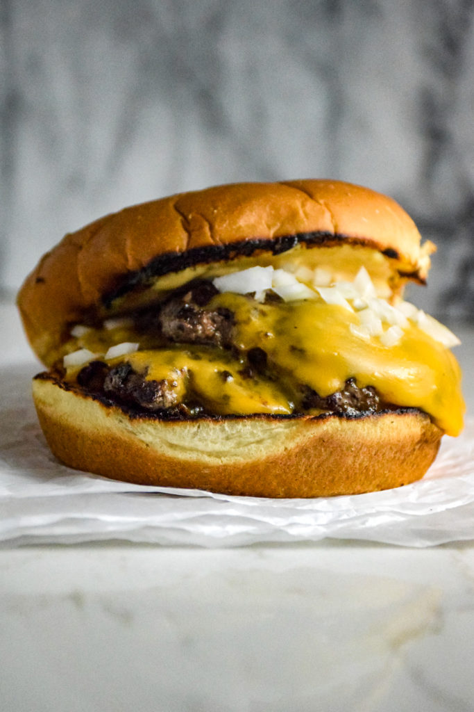 Burger with two thin patties on a bun with cheese and onions on a crumbled white paper on the grey marble background