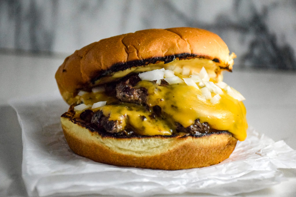 Burger with two thin patties on a bun with cheese and onions on a crumbled white paper on the grey marble background