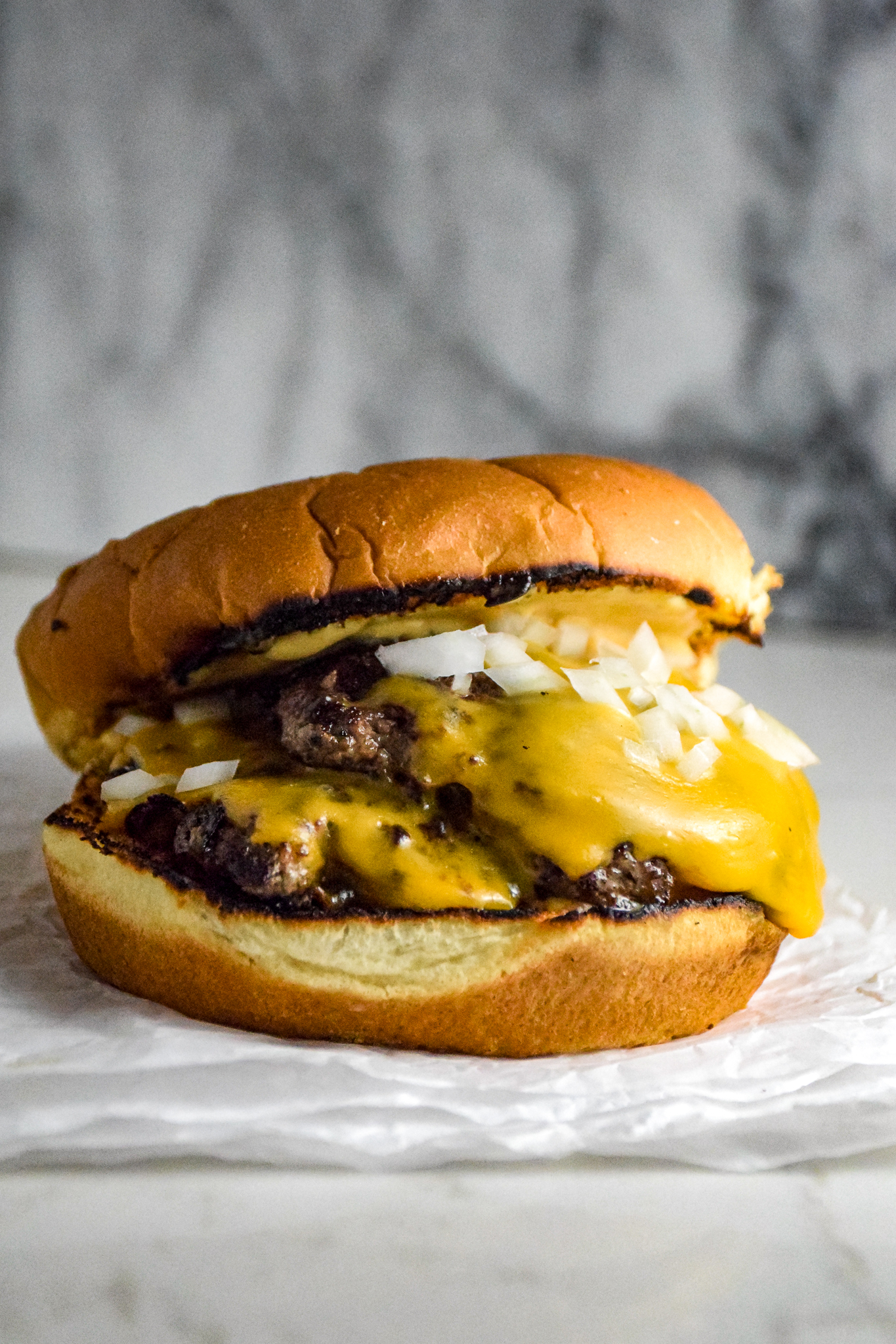 Burger with two thin patties on a bun with cheese and onions on a crumbled white paper on the grey marble background